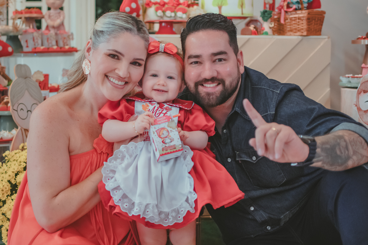 uma mulher de vestido vermelho e um homem de camisa azul segurando uma bebê de 1 aninho em frente uma decoração de chápeuzinho vermelho em uma festa de aniversário infantil.