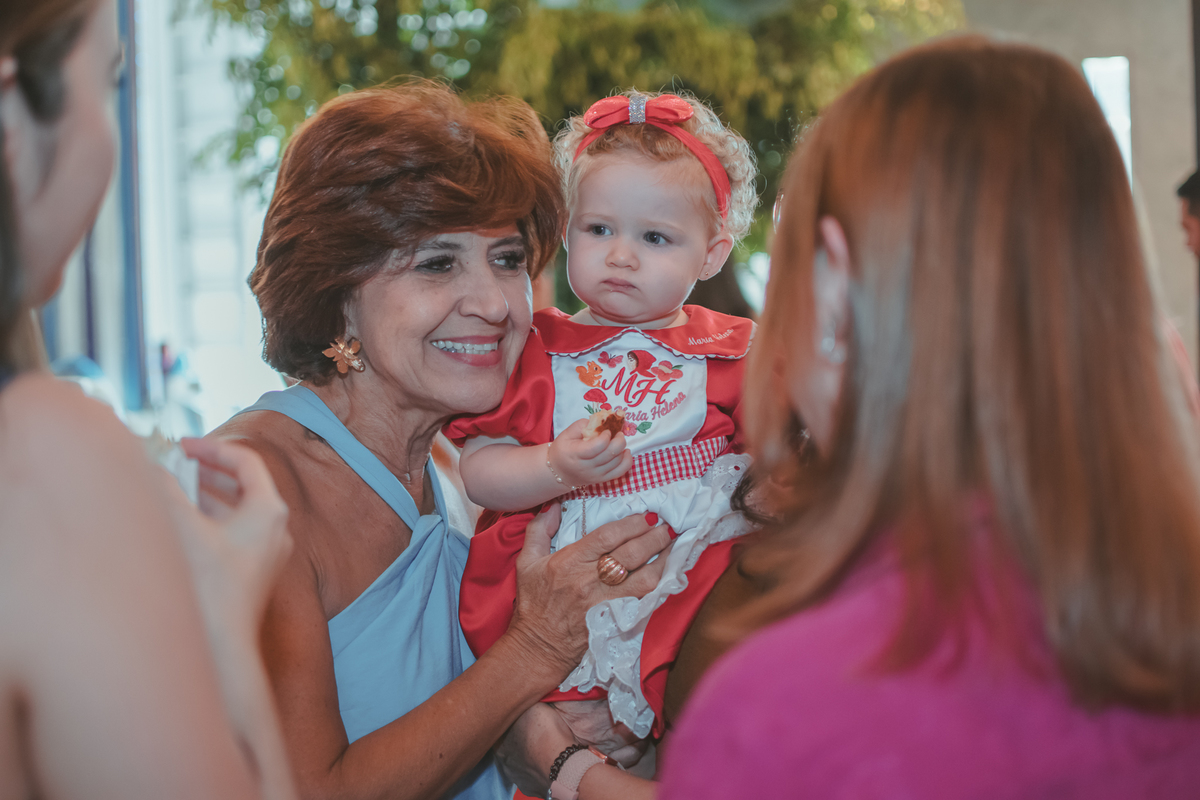 Uma senhora de idade sorrindo para uma mulher, enquanto abraça uma bebê na sua festa de aniversário de 1 ano.