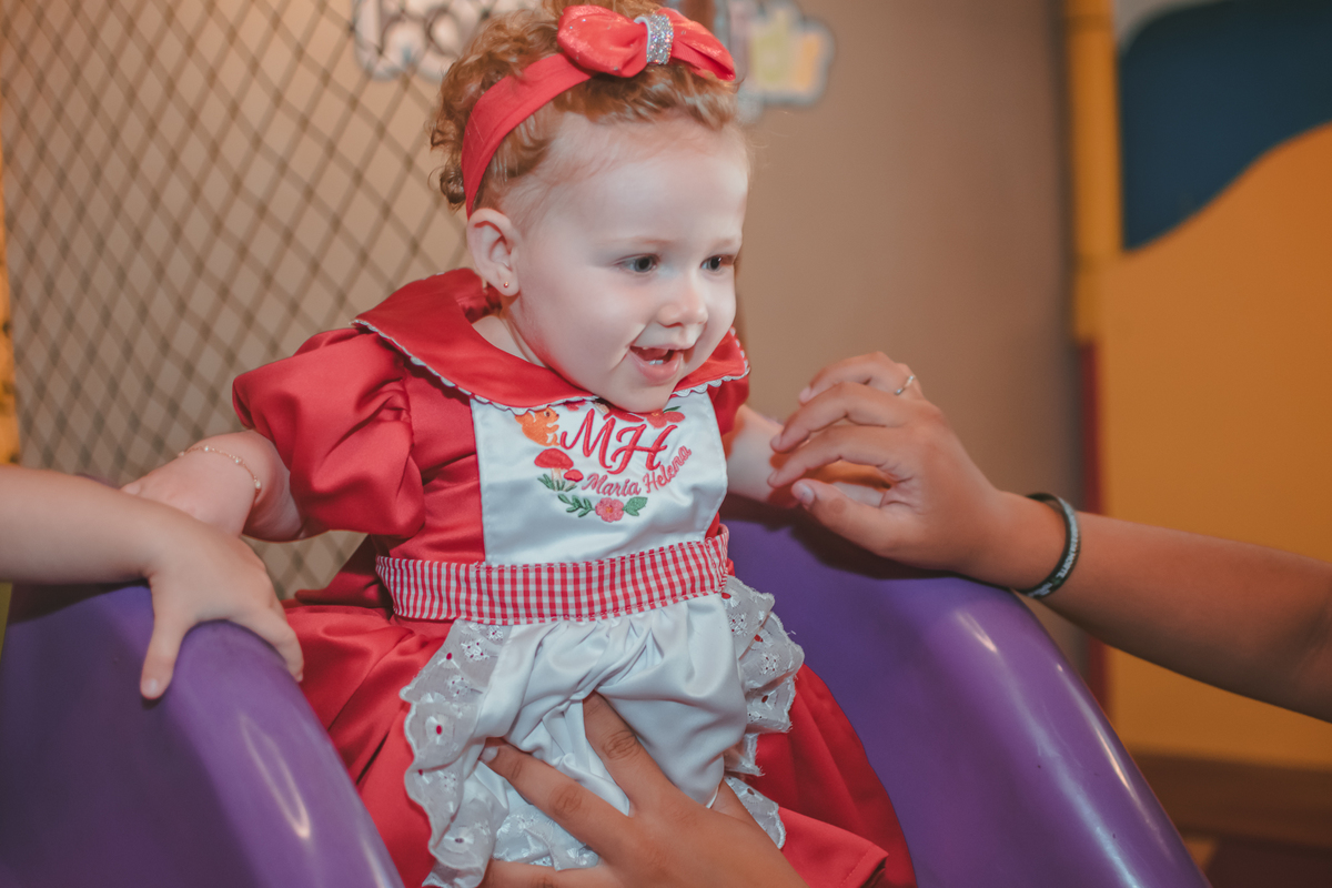Uma bebê de vestido vermelho e laço na cabeça, brincando na sua festa de aniversário de 1 ano.