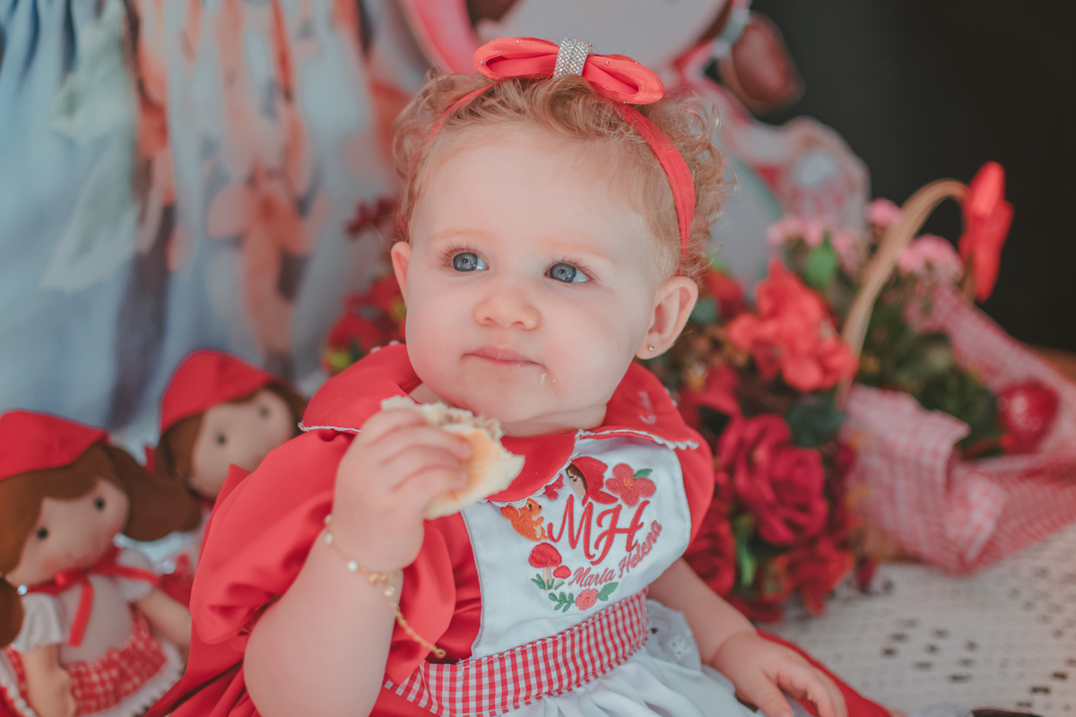 Uma bebê usando um vestido vermelho e comendo pão, durante sua festa de aniversário.