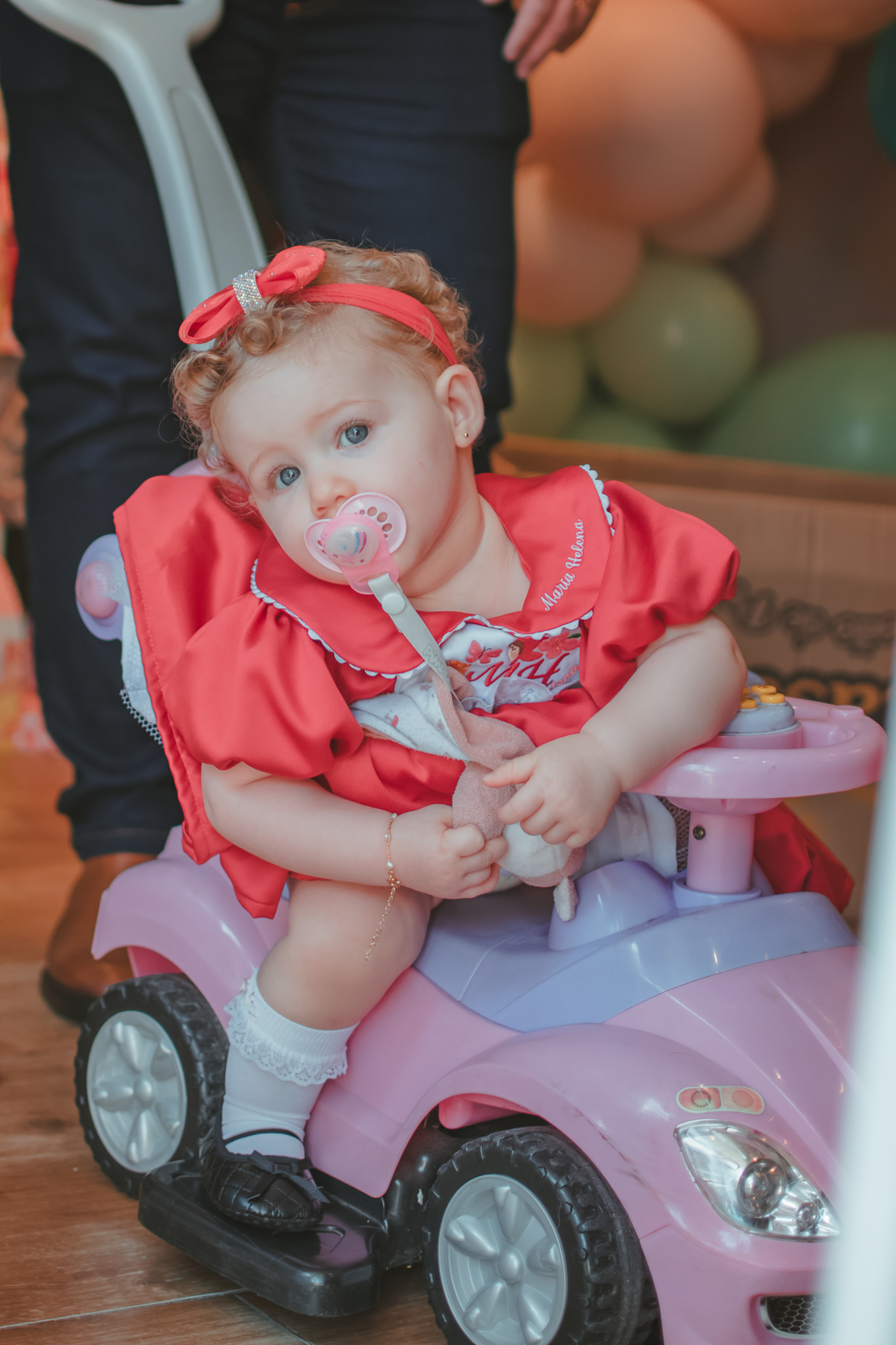 Uma bebê de 1 ano usando um vestido vermelho e sentada em um carinho de bebê, durante a sua festa de aniversário.