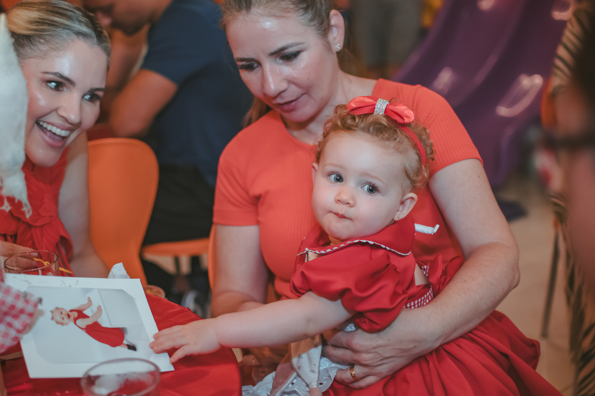 uma bebê, sentada no colo de uma mulher de vermelho e observando uma outra mulher, durante seu aniversário de 1 ano.