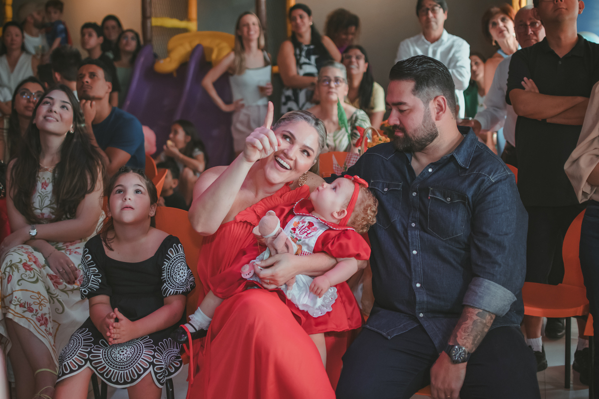 uma mulher com uma bebê no colo, um homem e uma criança ao lado, observando um vídeo de retrospectiva, durante uma festa de aniversário de 1 ano.