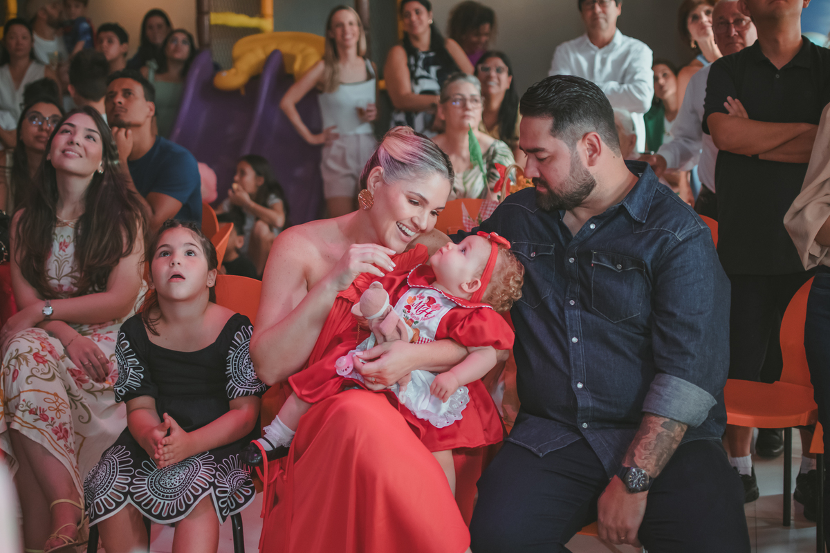 uma mulher com uma bebê no colo, um homem e uma criança ao lado, observando um vídeo de retrospectiva, durante uma festa de aniversário de 1 ano.