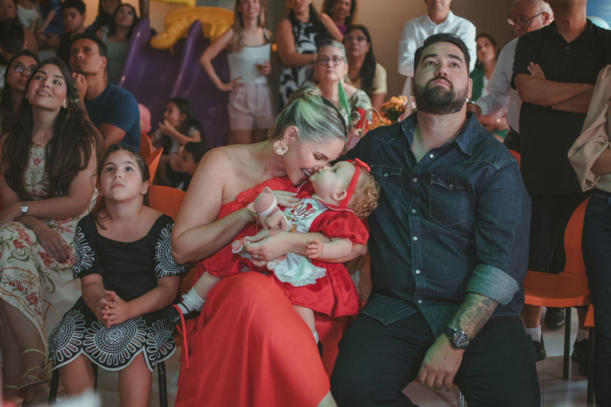 uma mulher com uma bebê no colo, um homem e uma criança ao lado, observando um vídeo de retrospectiva, durante uma festa de aniversário de 1 ano.