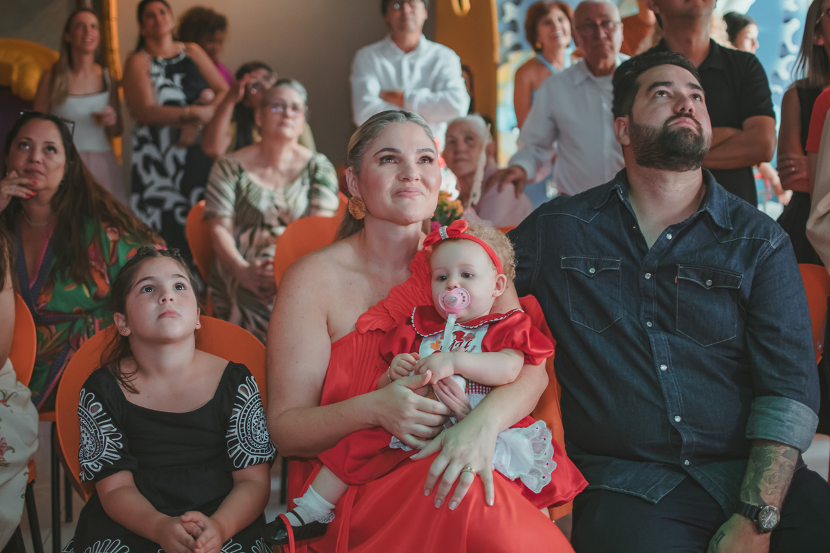 uma mulher com uma bebê no colo, um homem e uma criança ao lado, observando um vídeo de retrospectiva, durante uma festa de aniversário de 1 ano.