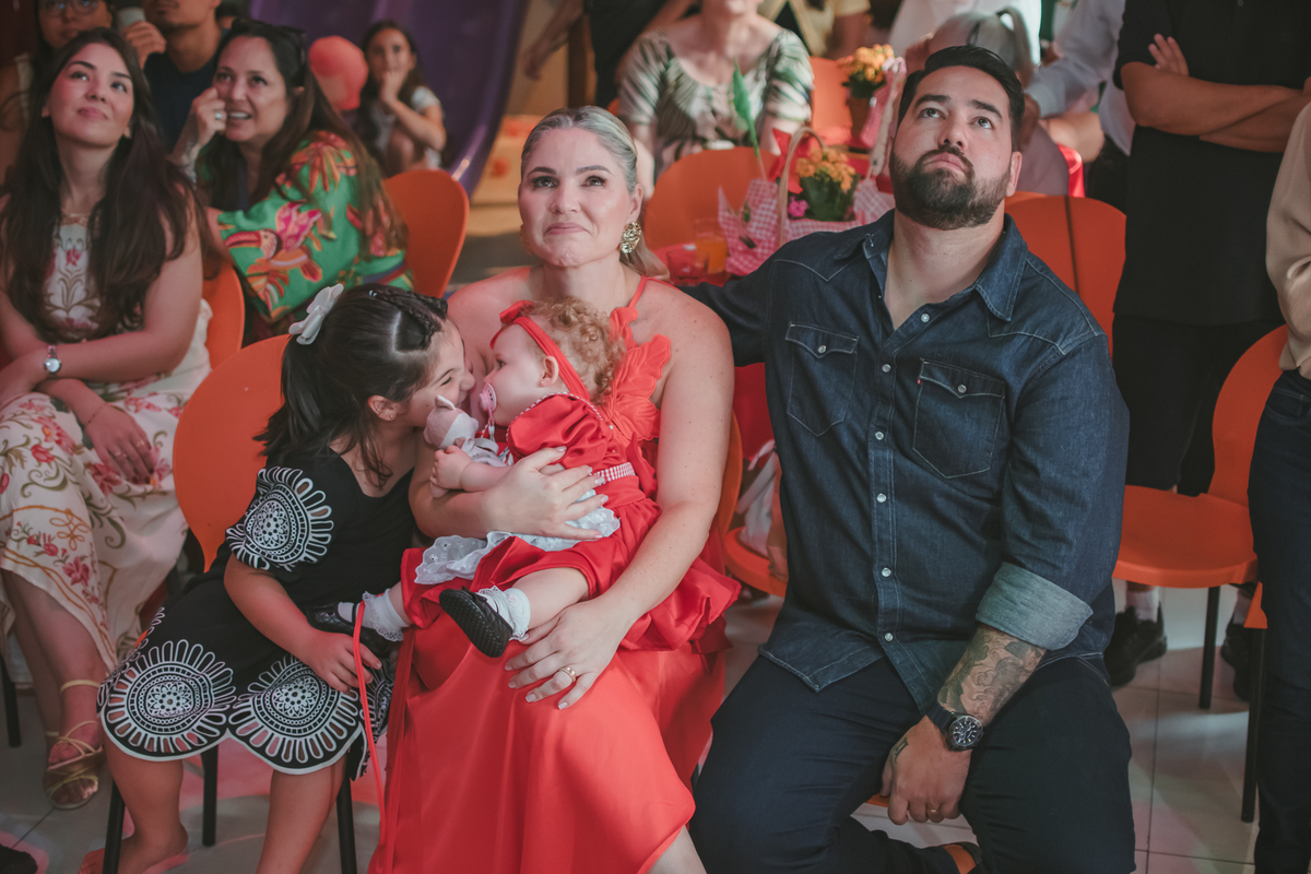 uma mulher com uma bebê no colo, um homem e uma criança ao lado, observando um vídeo de retrospectiva, durante uma festa de aniversário de 1 ano.