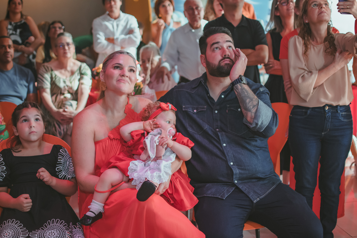 uma mulher com uma bebê no colo, um homem e uma criança ao lado, observando um vídeo de retrospectiva, durante uma festa de aniversário de 1 ano.