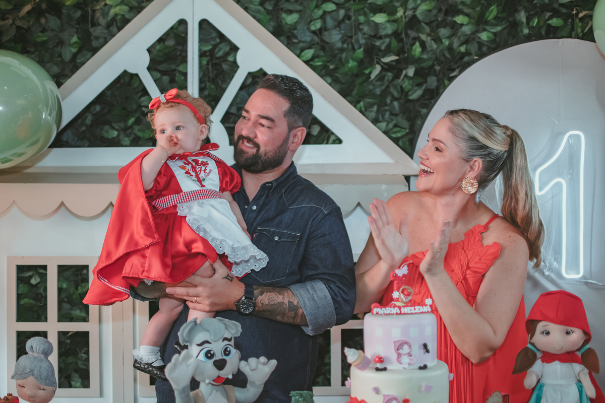 uma mulher de vermelho, um homem com camisa azul e uma bebê no colo, cantando parabéns em uma festa com decoração de Chapéuzinho Vermelho.