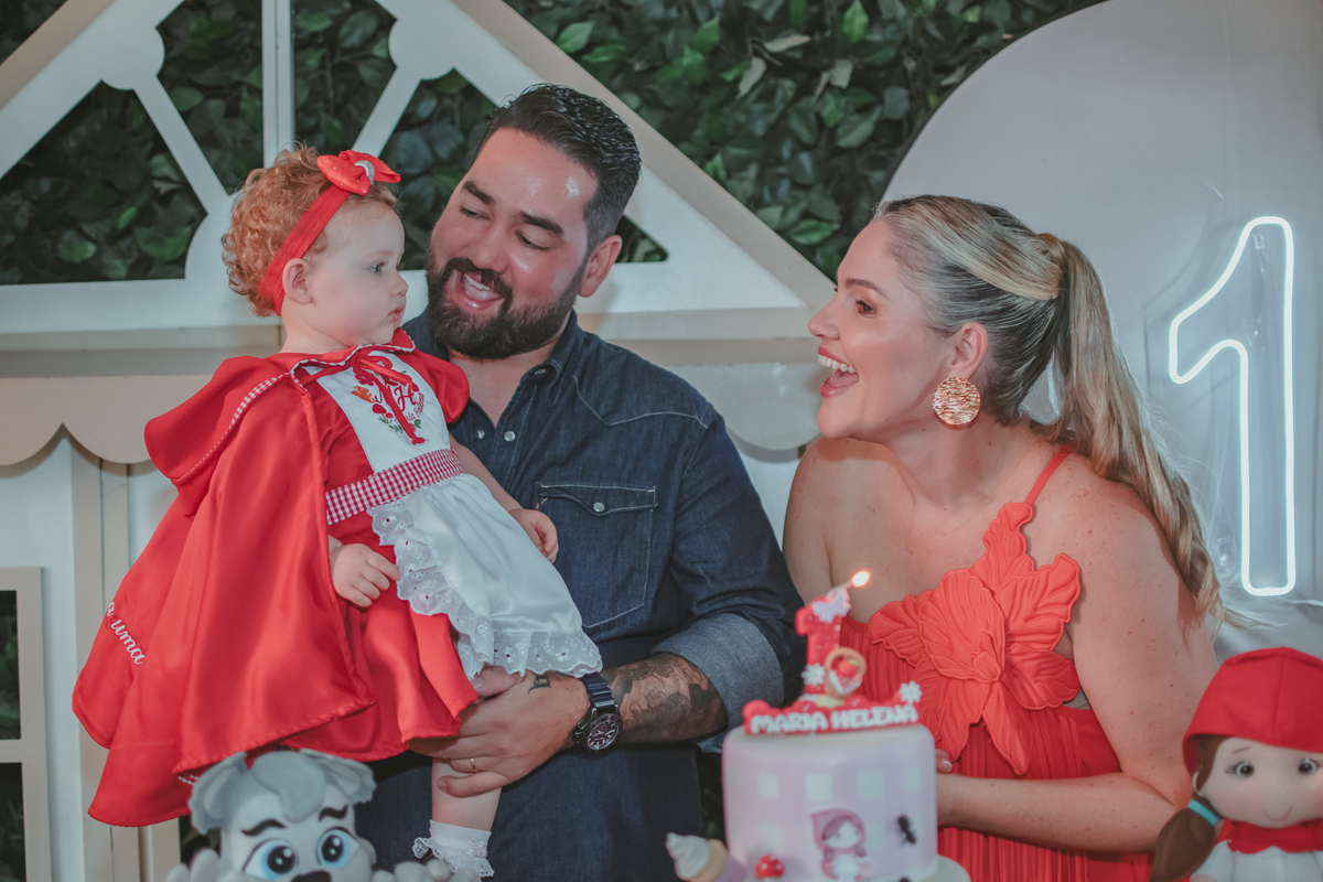 uma mulher de vermelho, um homem com camisa azul e uma bebê no colo, cantando parabéns em uma festa com decoração de Chapéuzinho Vermelho.