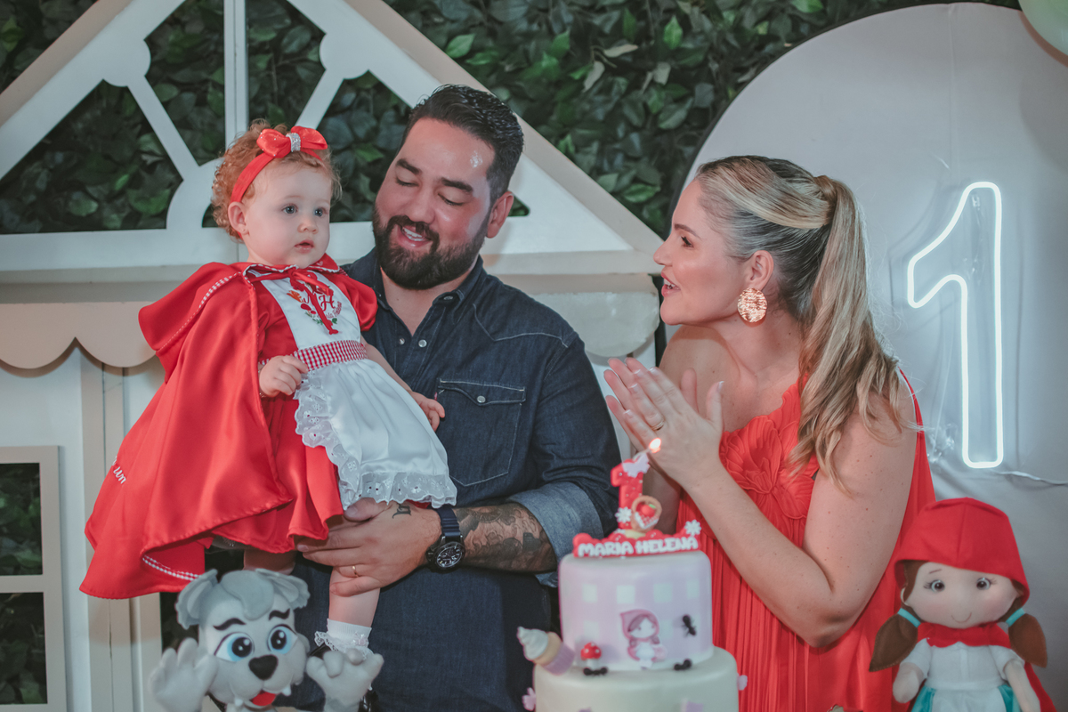 uma mulher de vermelho, um homem com camisa azul e uma bebê no colo, cantando parabéns em uma festa com decoração de Chapéuzinho Vermelho.