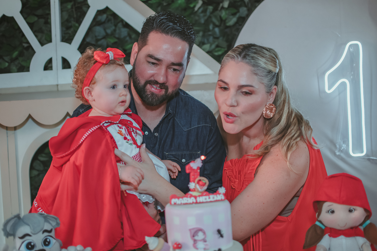 uma mulher de vermelho, um homem com camisa azul e uma bebê no colo, assoprando velhinha de 1 ano em uma festa com decoração de Chapéuzinho Vermelho.