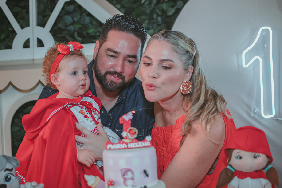 uma mulher de vermelho, um homem com camisa azul e uma bebê no colo, assoprando velhinha de 1 ano em uma festa com decoração de Chapéuzinho Vermelho.