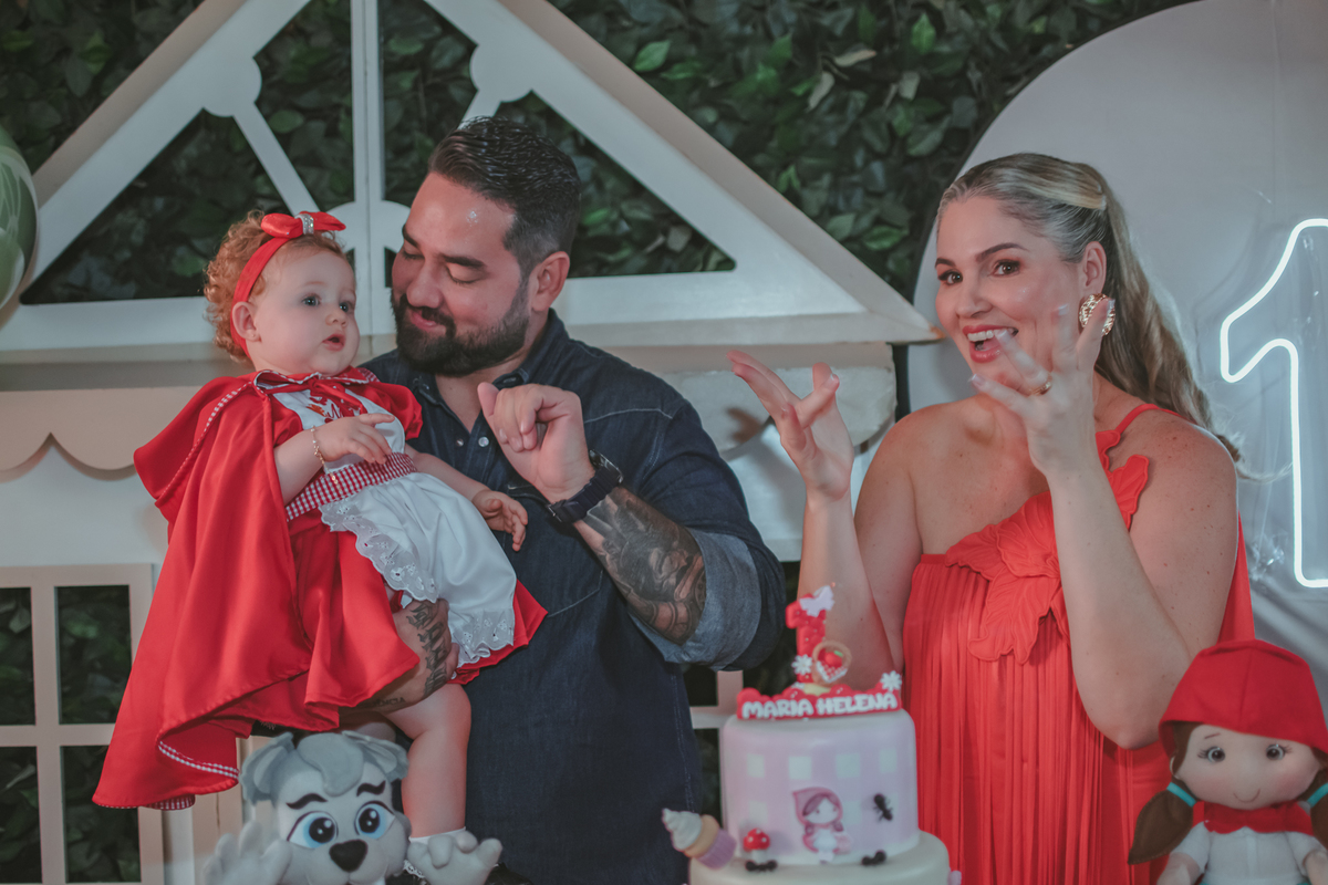 uma mulher de vermelho, um homem com camisa azul e uma bebê no colo, cantando parabéns em uma festa com decoração de Chapéuzinho Vermelho.