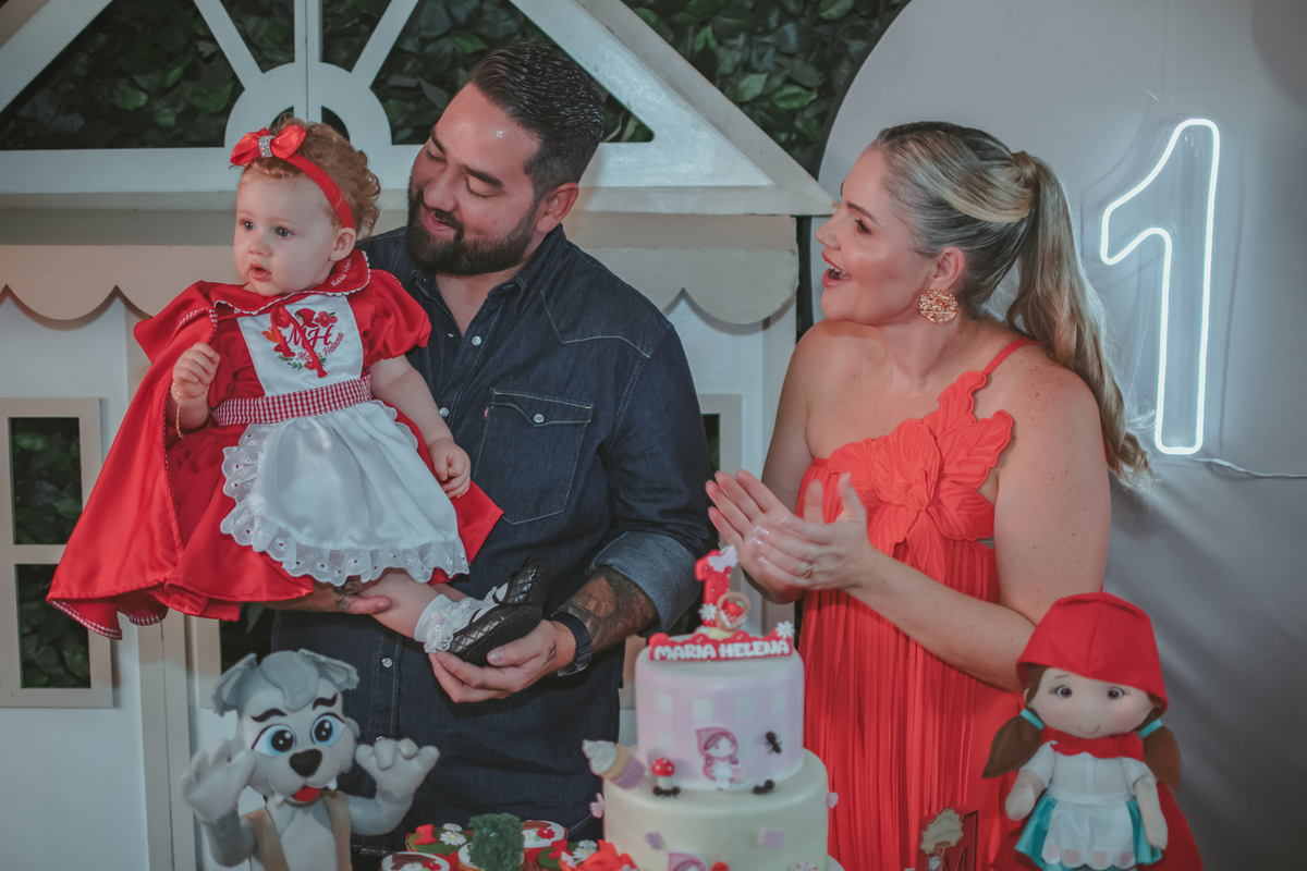 uma mulher de vermelho, um homem com camisa azul e uma bebê no colo, cantando parabéns em uma festa com decoração de Chapéuzinho Vermelho.