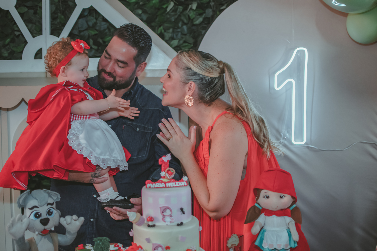 uma mulher de vermelho, um homem com camisa azul e uma bebê no colo, cantando parabéns em uma festa com decoração de Chapéuzinho Vermelho.