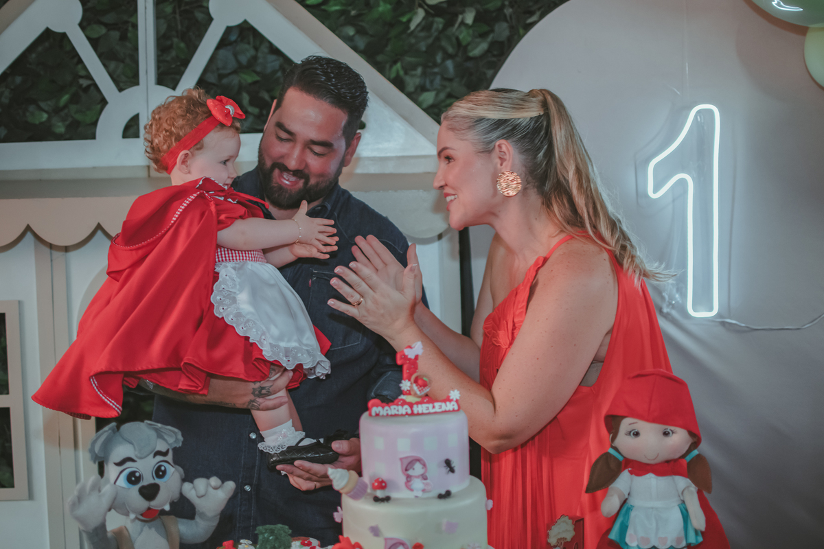 uma mulher de vermelho, um homem com camisa azul e uma bebê no colo, cantando parabéns em uma festa com decoração de Chapéuzinho Vermelho.