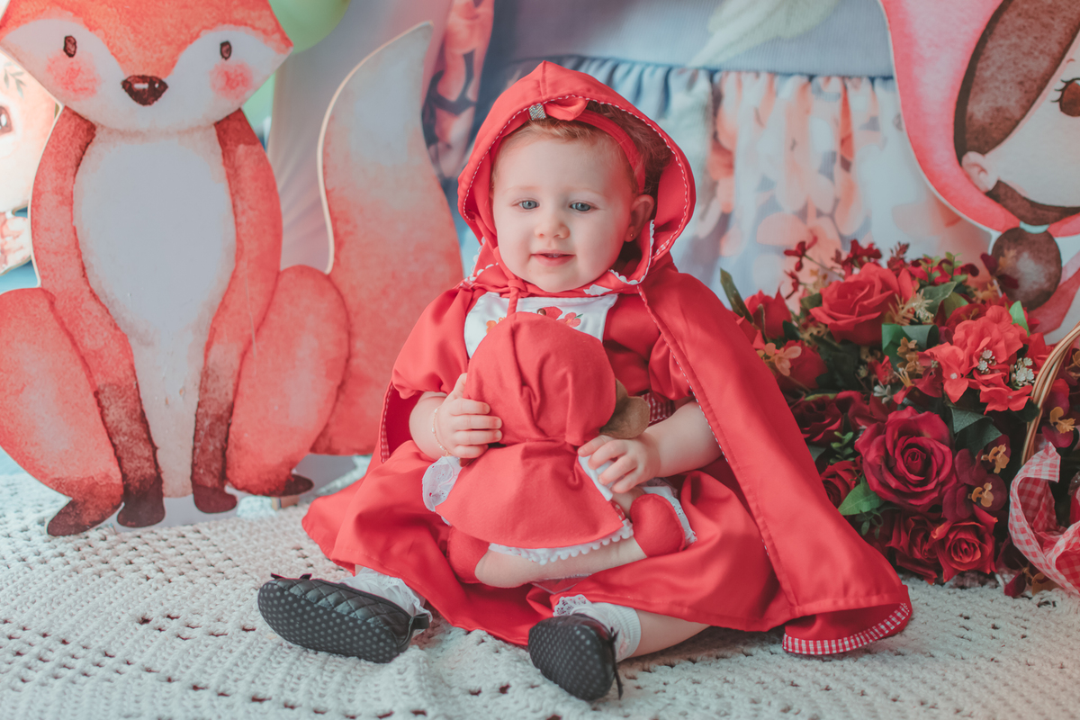 uma bebê de 1 ano usando uma capa Vermelha e brincando com uma boneca de Chapéuzinho Vermelho em sua festa de aniversário.
