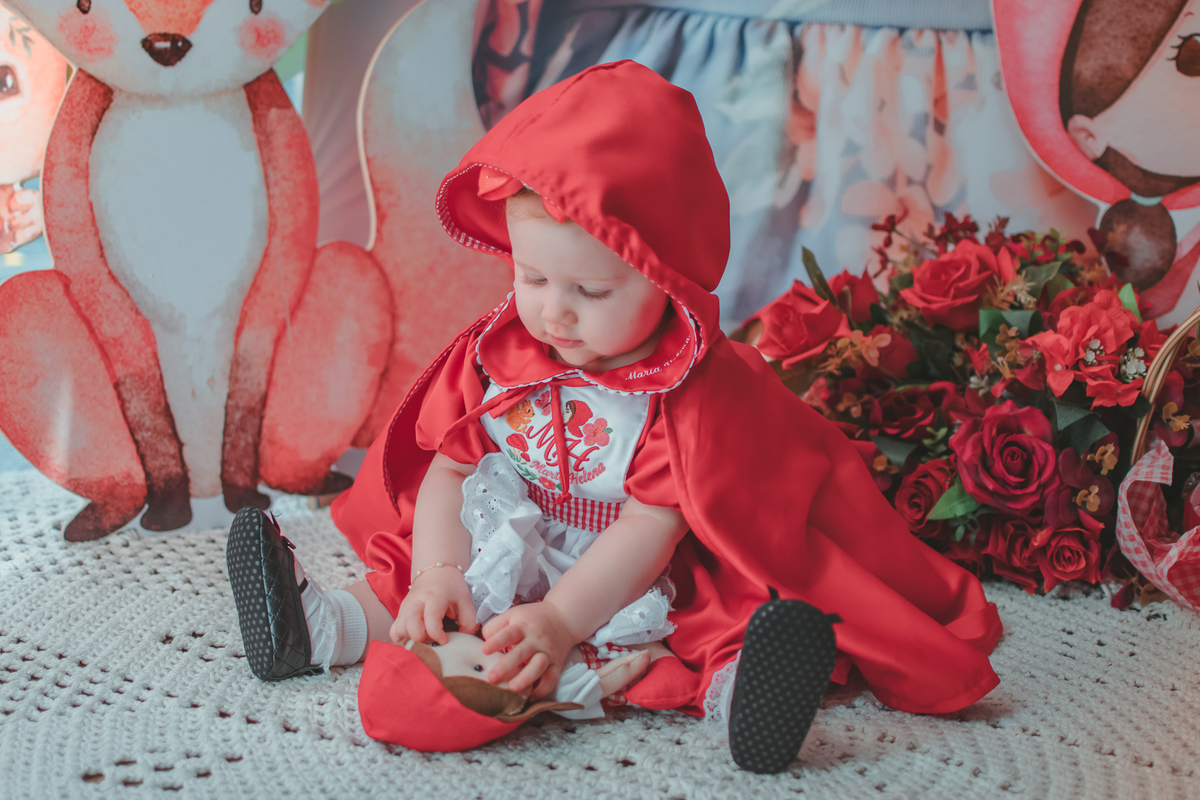 uma bebê de 1 ano usando uma capa Vermelha e brincando com uma boneca de Chapéuzinho Vermelho em sua festa de aniversário.