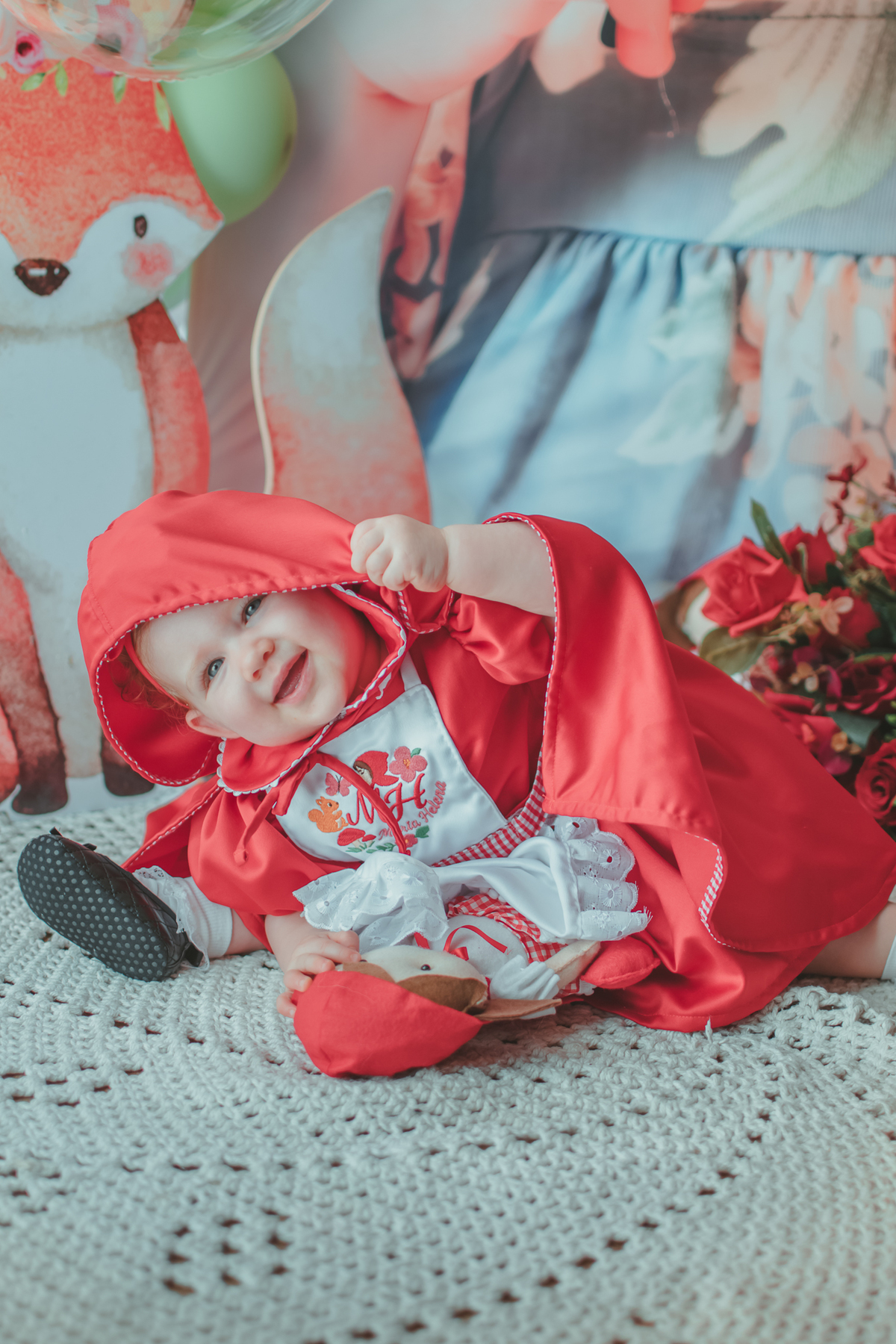uma bebê de 1 ano usando uma capa Vermelha e brincando com uma boneca de Chapéuzinho Vermelho em sua festa de aniversário.