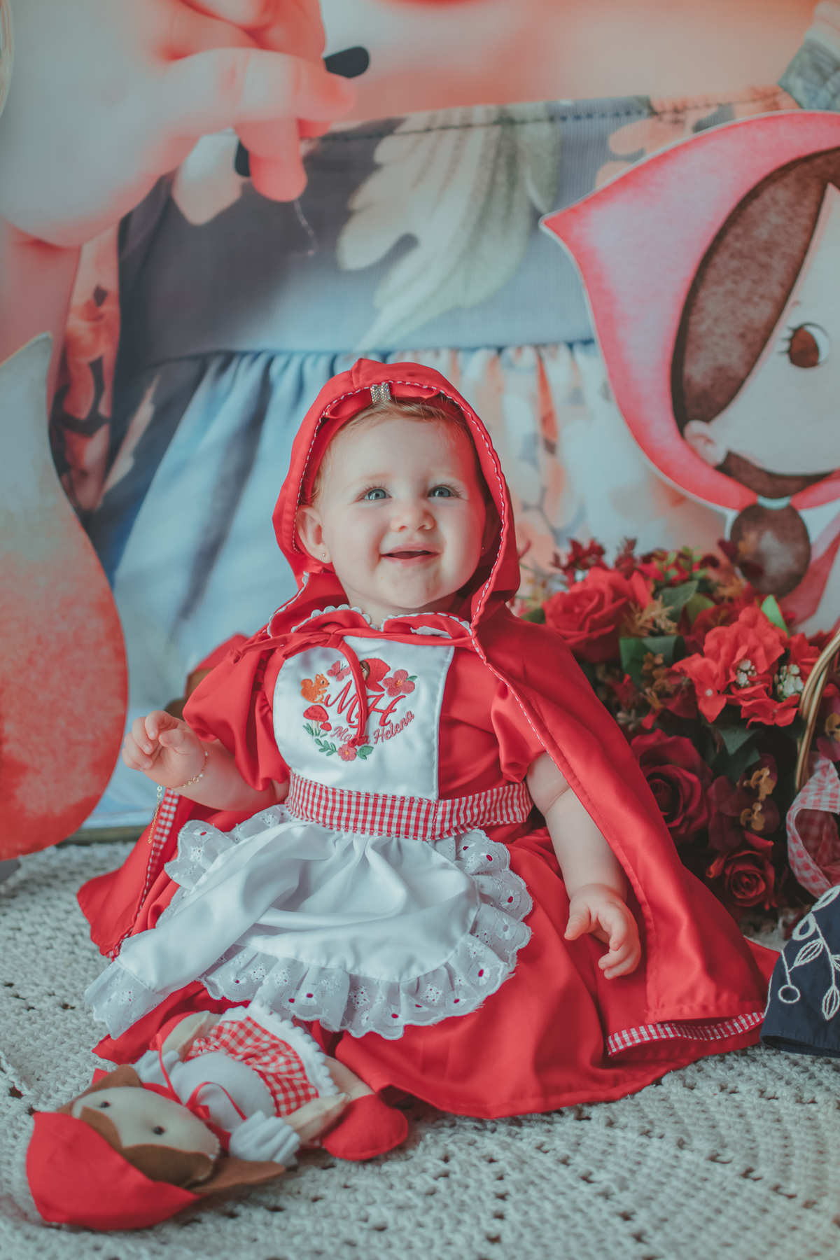 uma bebê de 1 ano usandouma capa Vermelha e brincando com uma boneca de Chapéuzinho Vermelho em sua festa de aniversário.