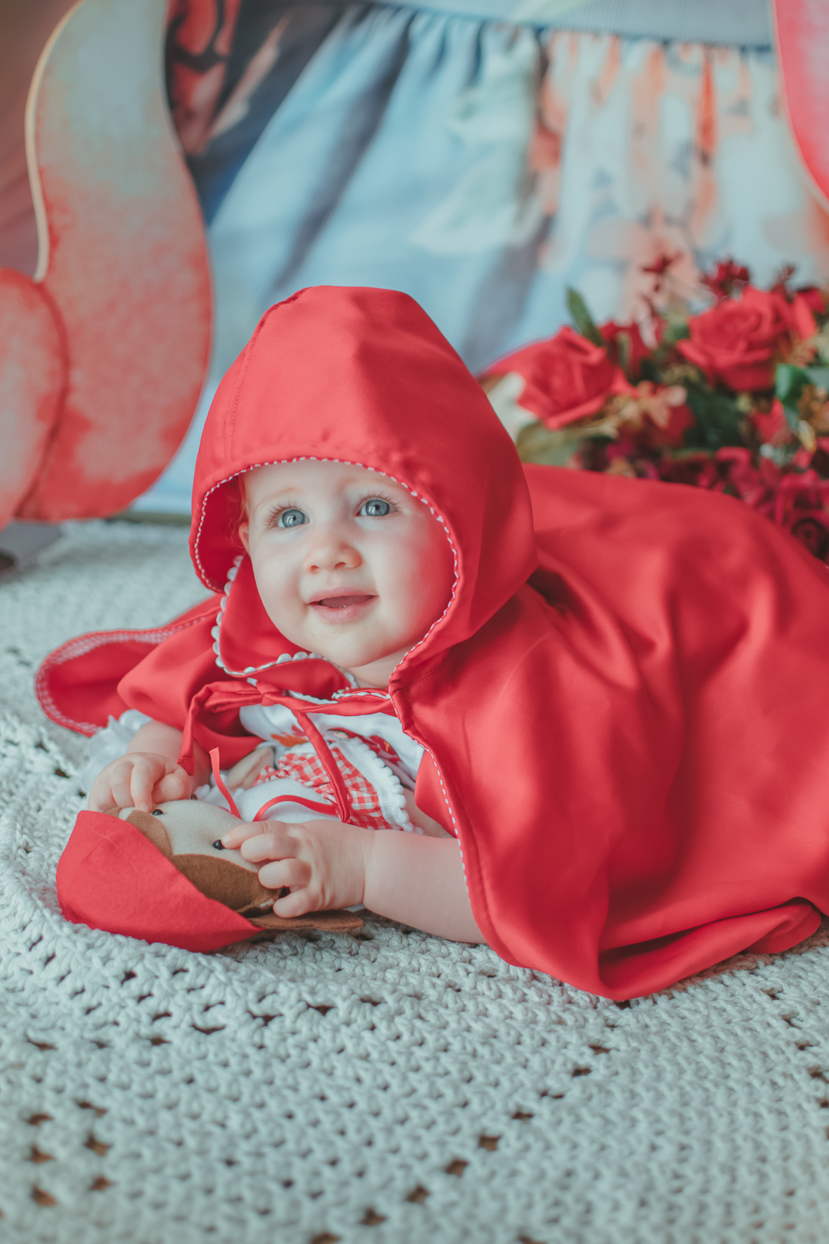 uma bebê de 1 ano usandouma capa Vermelha e brincando com uma boneca de Chapéuzinho Vermelho em sua festa de aniversário.