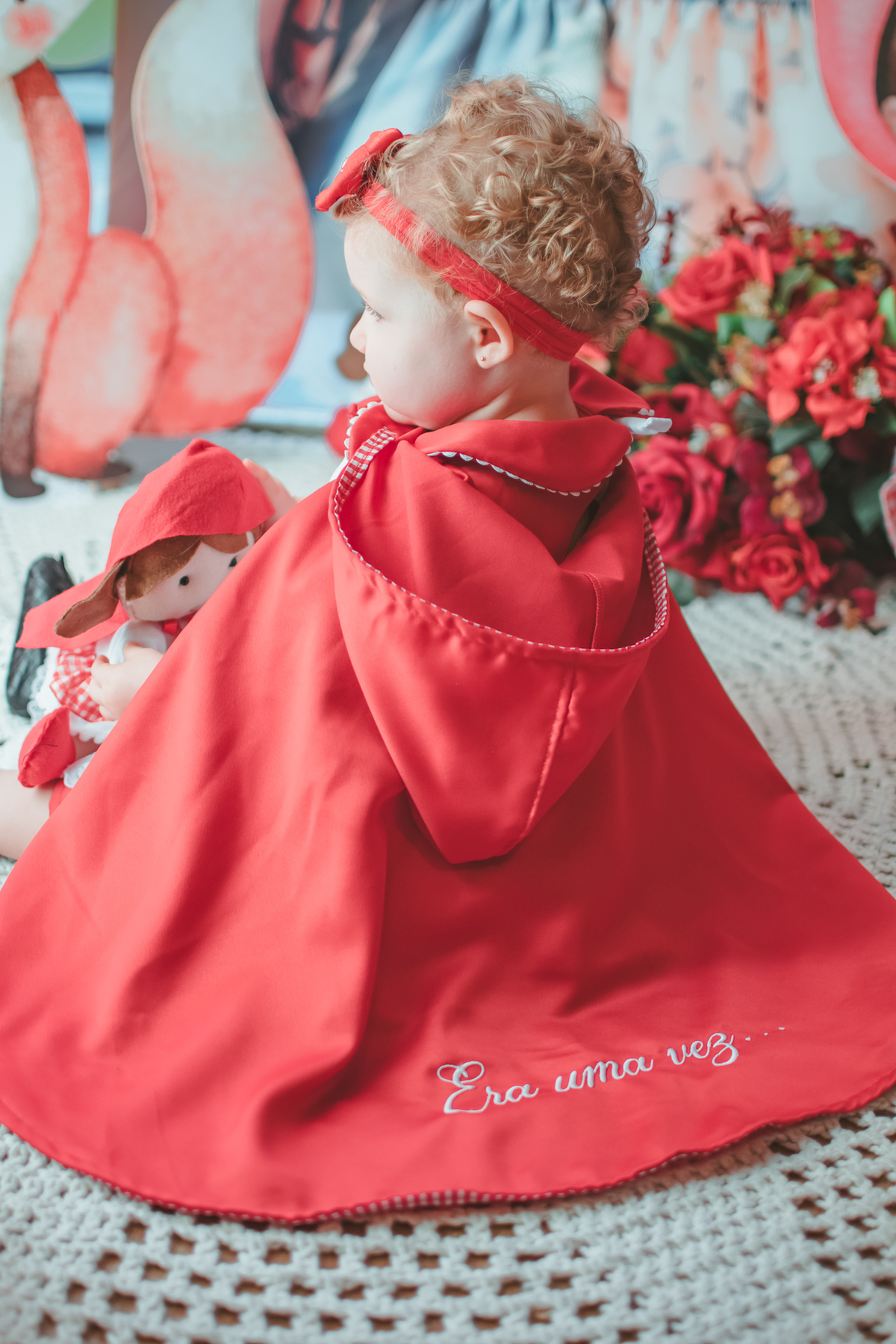 uma bebê de 1 ano usando uma capa Vermelha e brincando com uma boneca de Chapéuzinho Vermelho em sua festa de aniversário.