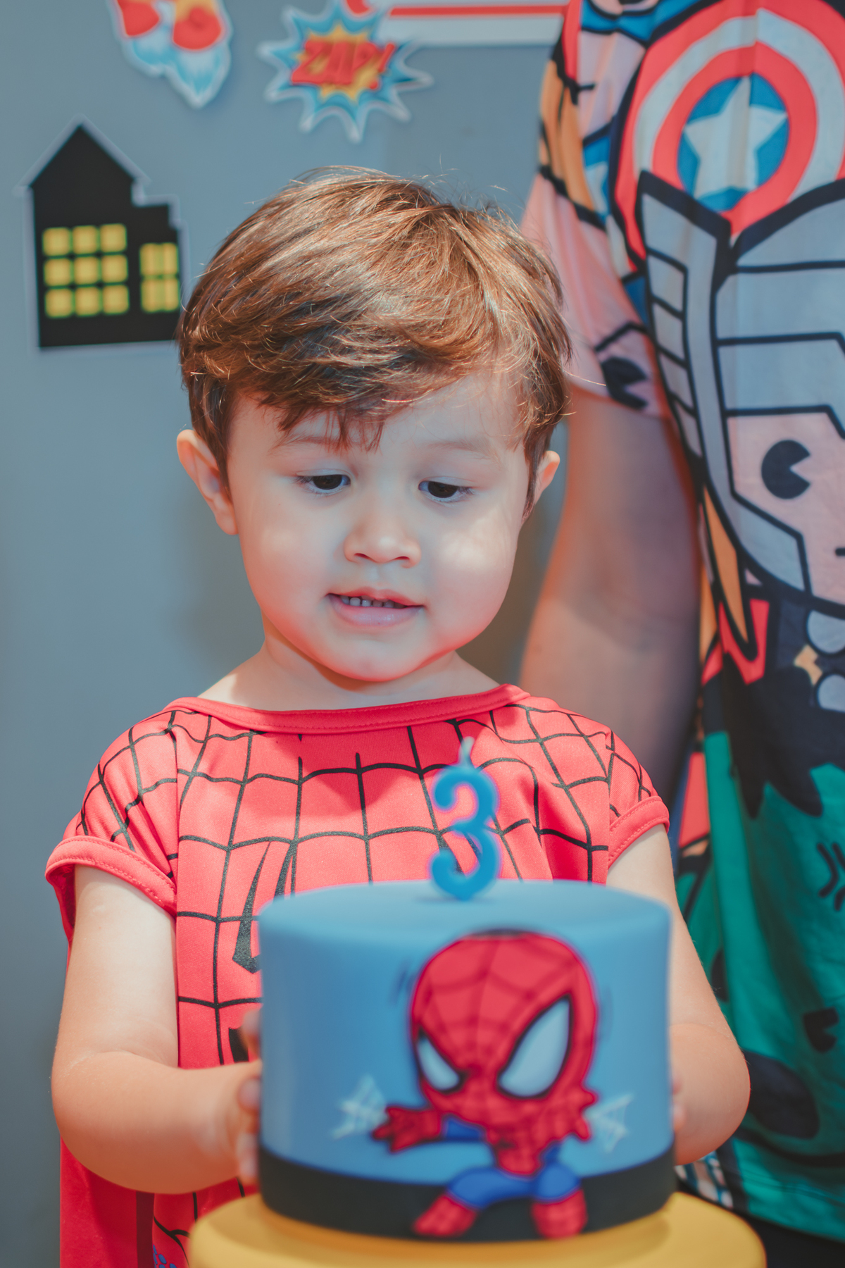Um menino vestido com fantasia de homem aranha em frente a um bolo de aniversário com uma vela de 3 anos.