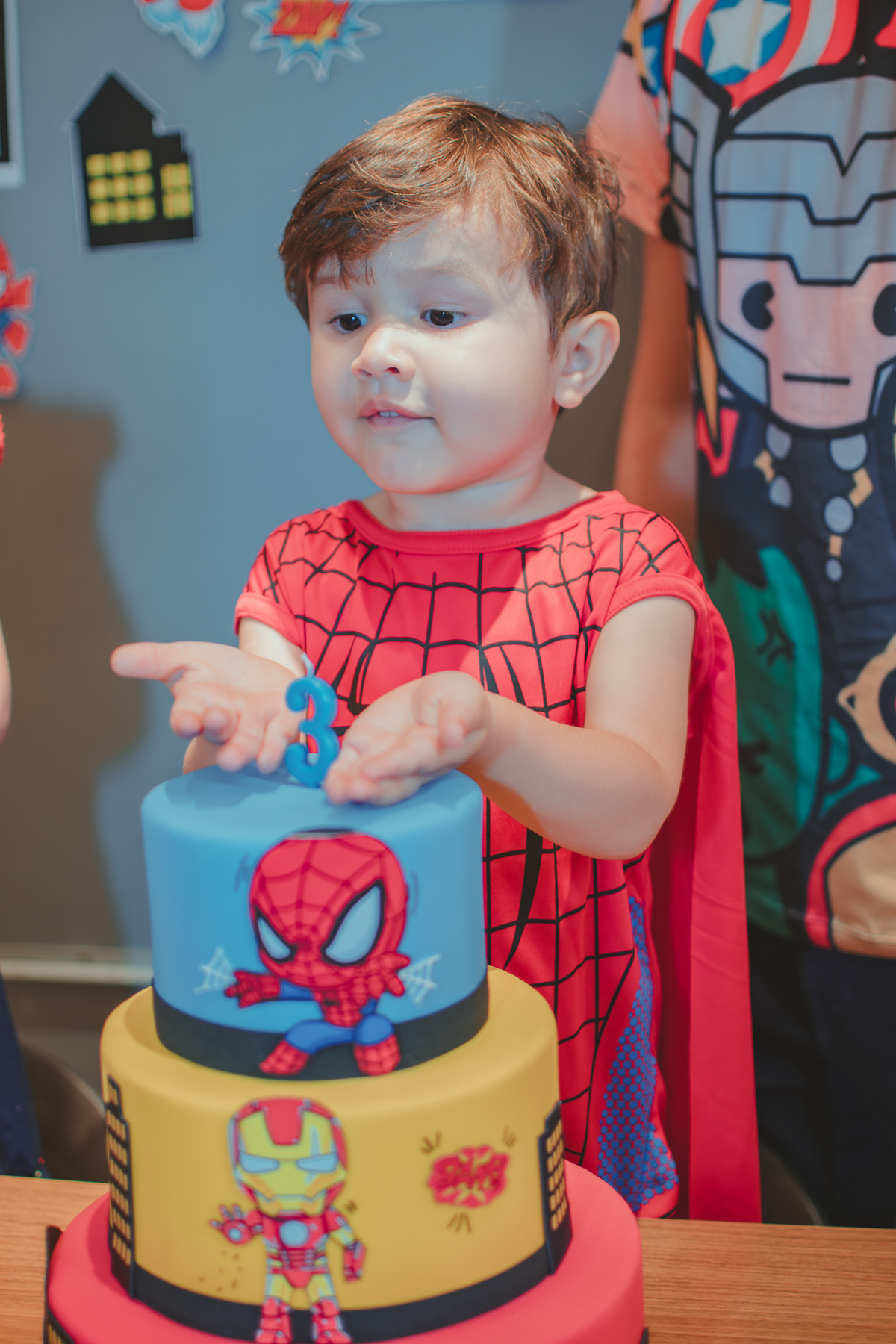 Um menino vestido com fantasia de homem aranha apontando a um bolo de aniversário com uma vela de 3 anos.