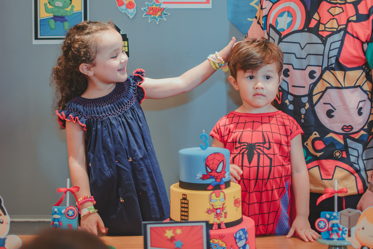 Uma menina com vestido azul acariciando os cabelos do seu irmão que está vestido com fantasia de homem aranha durante o parabéns de uma festa de aniversário.