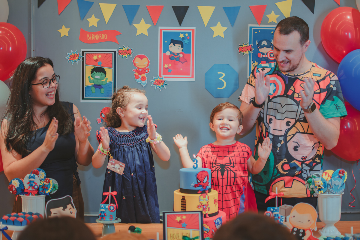 Uma familia reunida em frente uma mesa de bolo cantando parabéns para um menino vestido com fantasia de Homem Aranha.