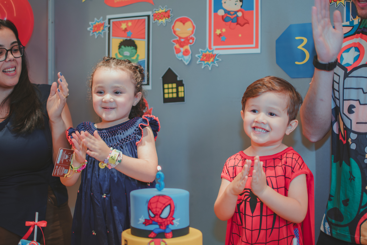 Uma familia reunida em frente uma mesa de bolo cantando parabéns para um menino vestido com fantasia de Homem Aranha.
