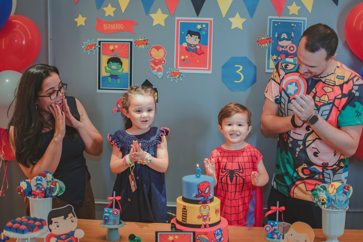 Uma familia reunida em frente uma mesa de bolo cantando parabéns para um menino vestido com fantasia de Homem Aranha.