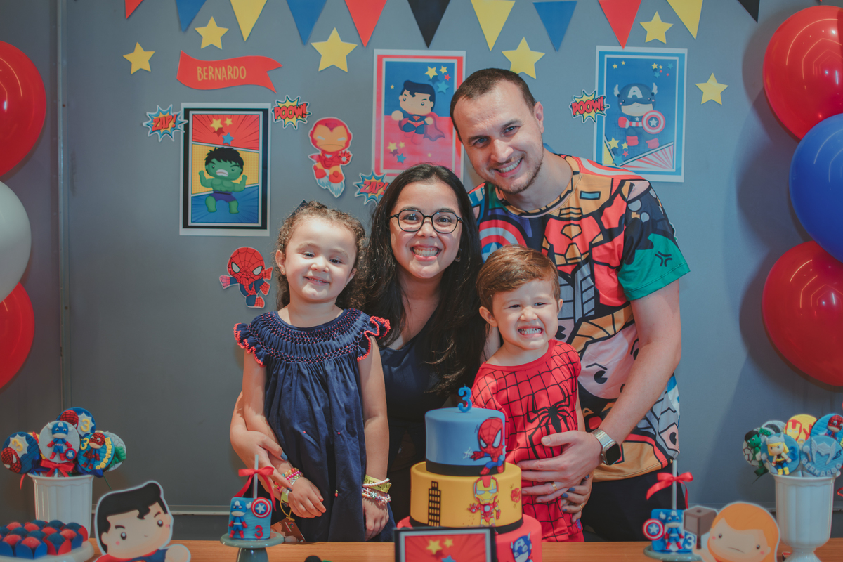 Uma familia  com duas crianças, juntos em frente uma mesa de bolo decorada com tema de super Heróis, durante uma festa de aniversário.