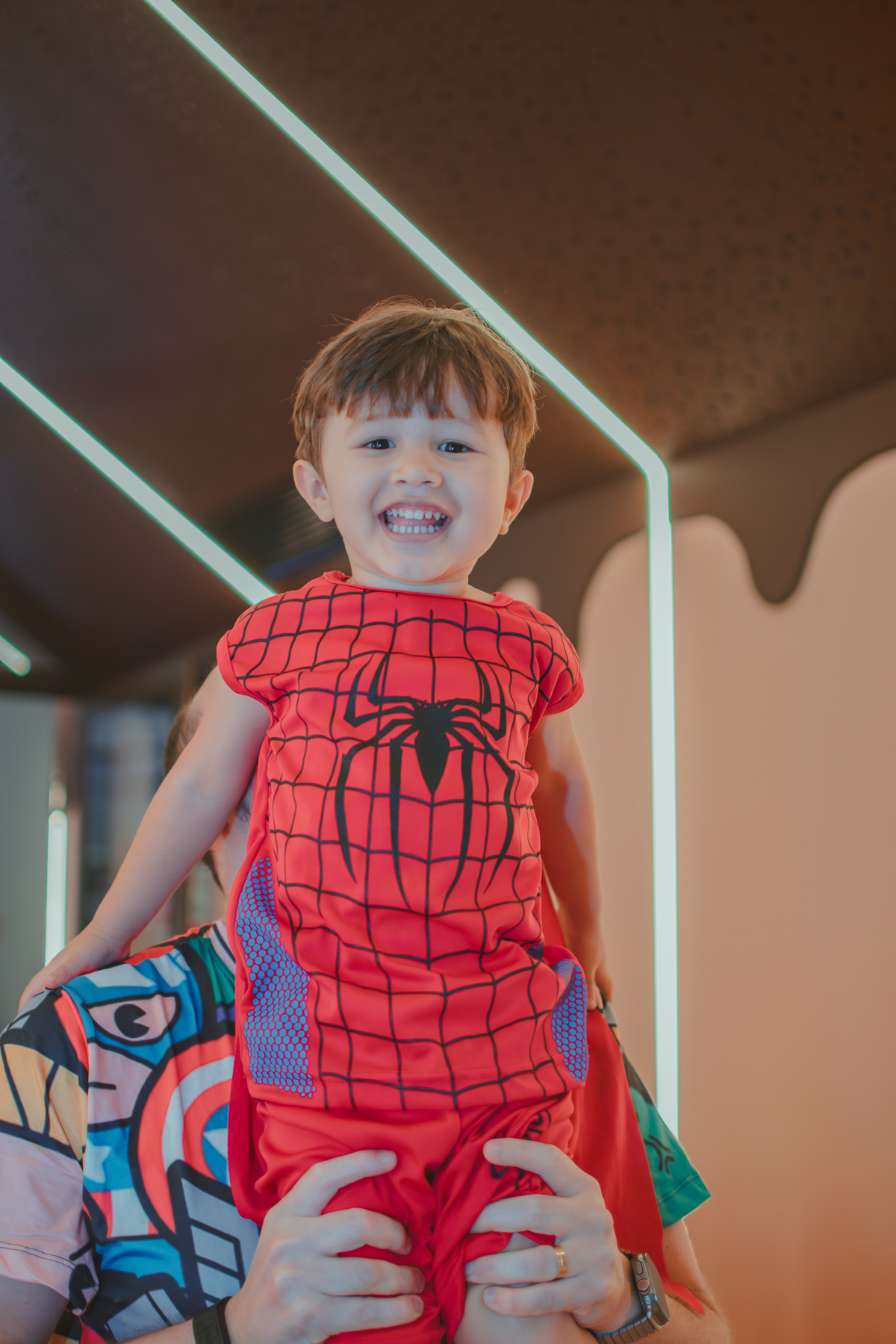 Um menino vestido de Homem Aranha, sorrindo enquanto seu pai o suspende no ar. 