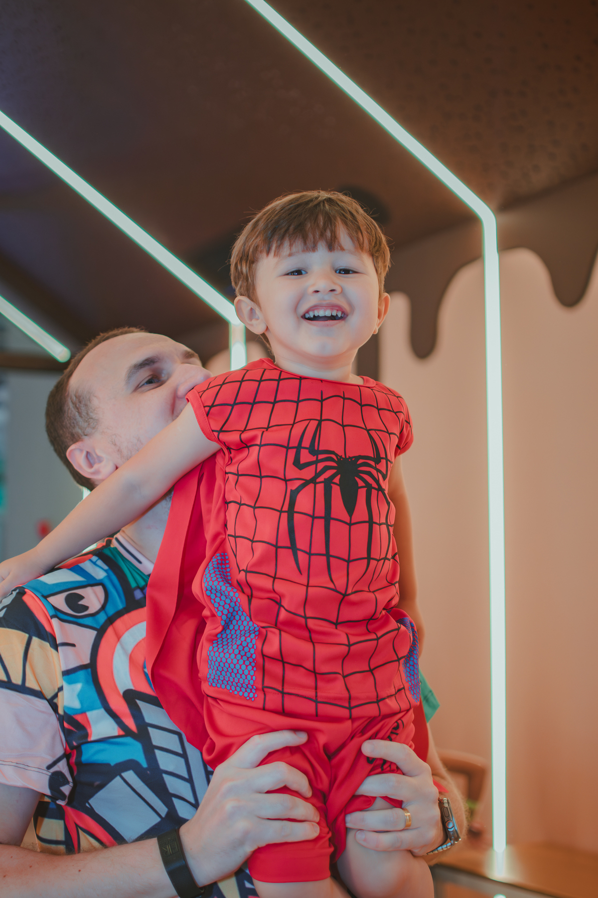 Um menino vestido de Homem Aranha, sorrindo enquanto seu pai o suspende no ar. 