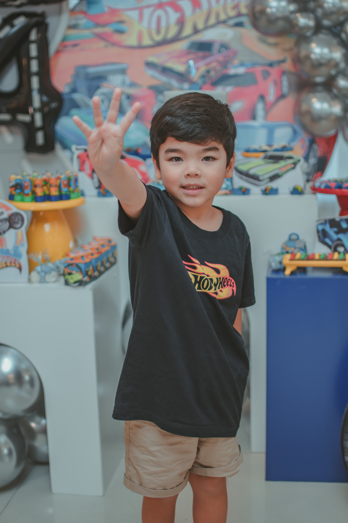 um menino vestido com camisa preto, em frente a decoração do seu aniversário fazendo o número 4 com a mão.
