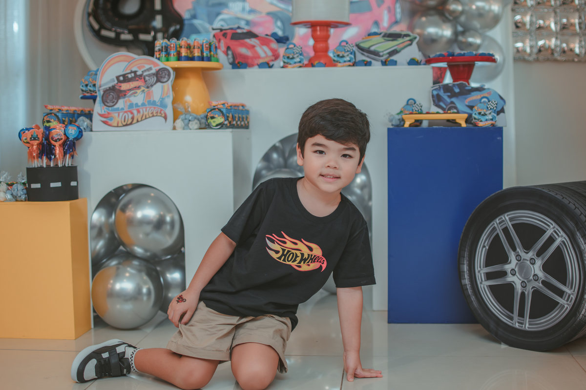 Um menino sentado em frente a decoração com tema Hot Wheels, durante sua festa de aniversário de 4 anos.