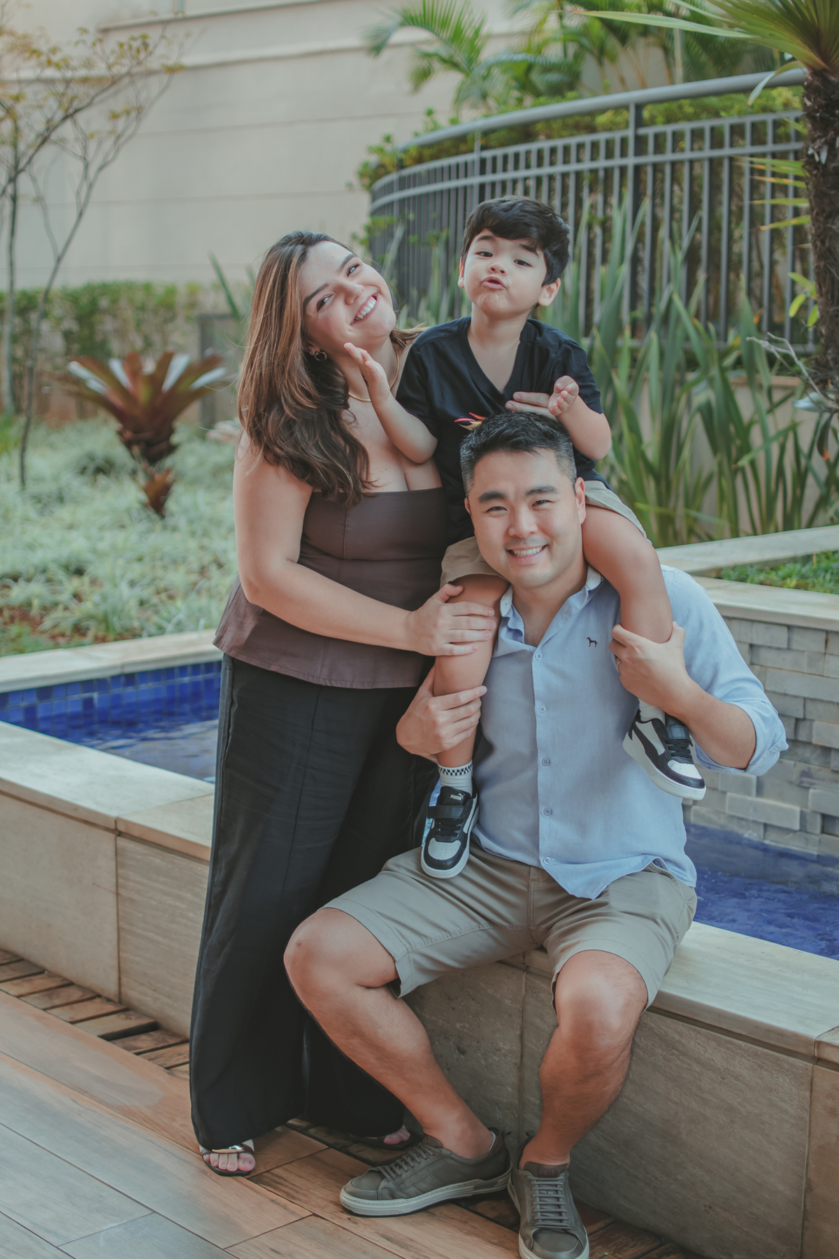 um homem, com um menino vestido de camiseta preta em seu ombro e a mãe da criança ao lado, durante um festa de aniversário infantil.