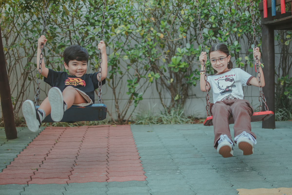 um menino de camiseta preta e uma menina de camiseta branca, brincando no balanço durante uma festa de aniversário infantil.