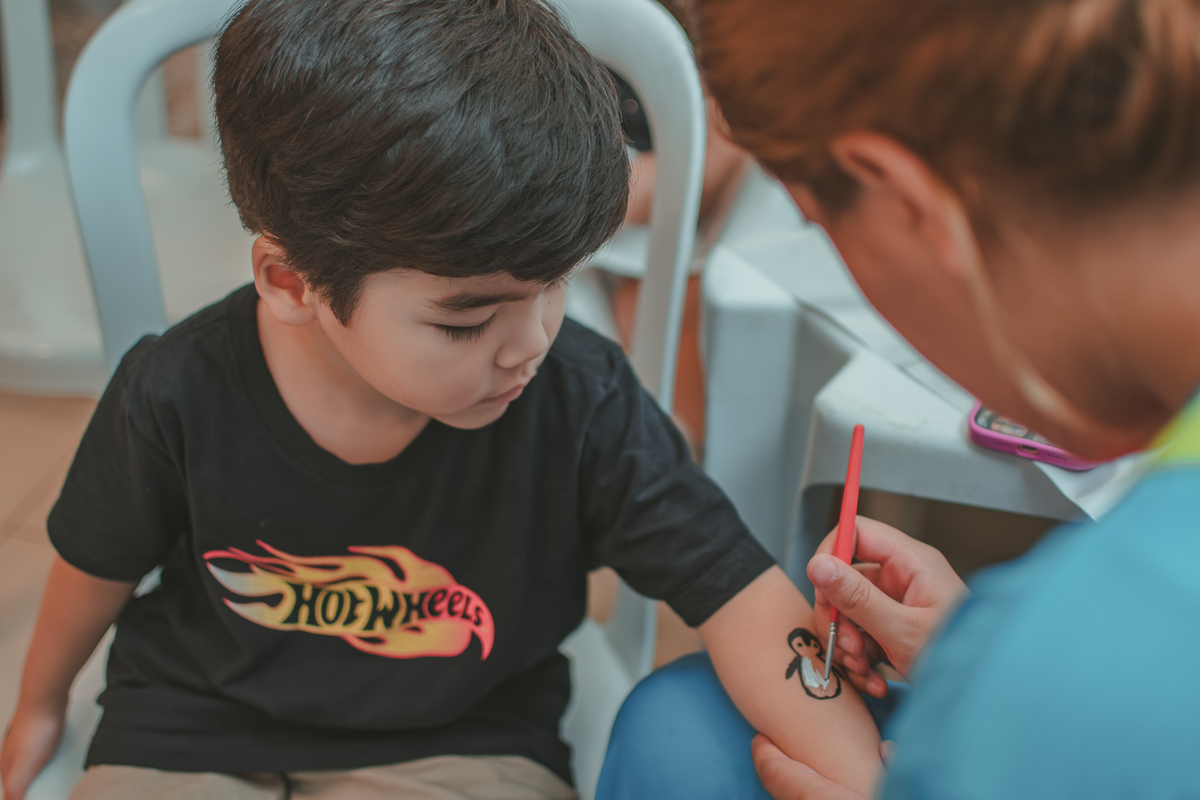Um menino de camiseta preta, pintando o desenho de um pinguim no branço, durante sua festa de aniverrsário de 4 anos.
