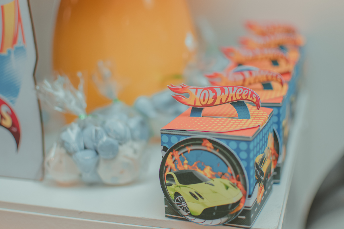Caixinha decorada com um carro vermelho e a escrita Hot Wheels em cima. Ao lado suspiros branco de azul dentro de uma saquinho transparente, compondo da decoração de aniversário de menino.