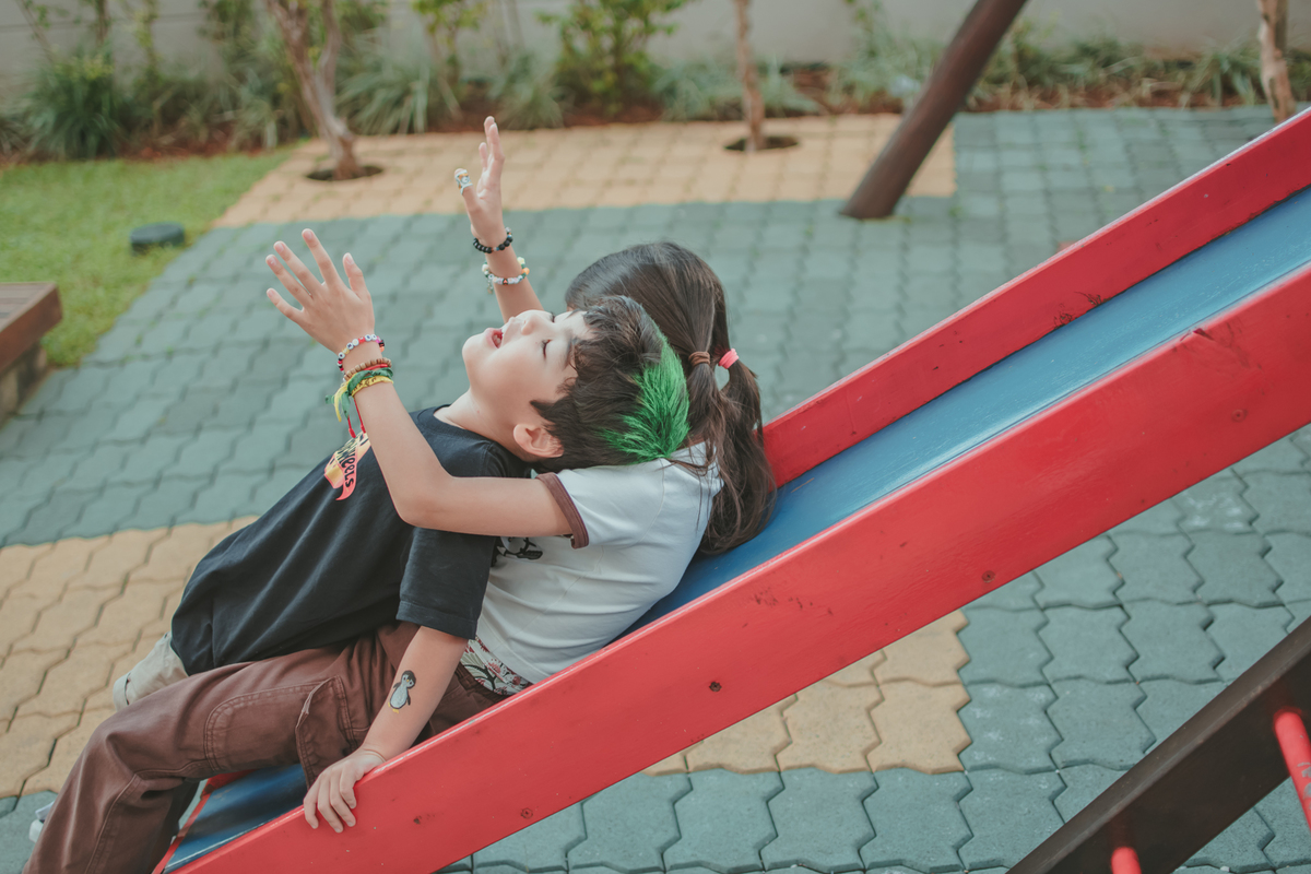 um menino de camiseta preta e uma menina de camiseta branca brincando de descer no escorregador, durante sua festa de aniversário.