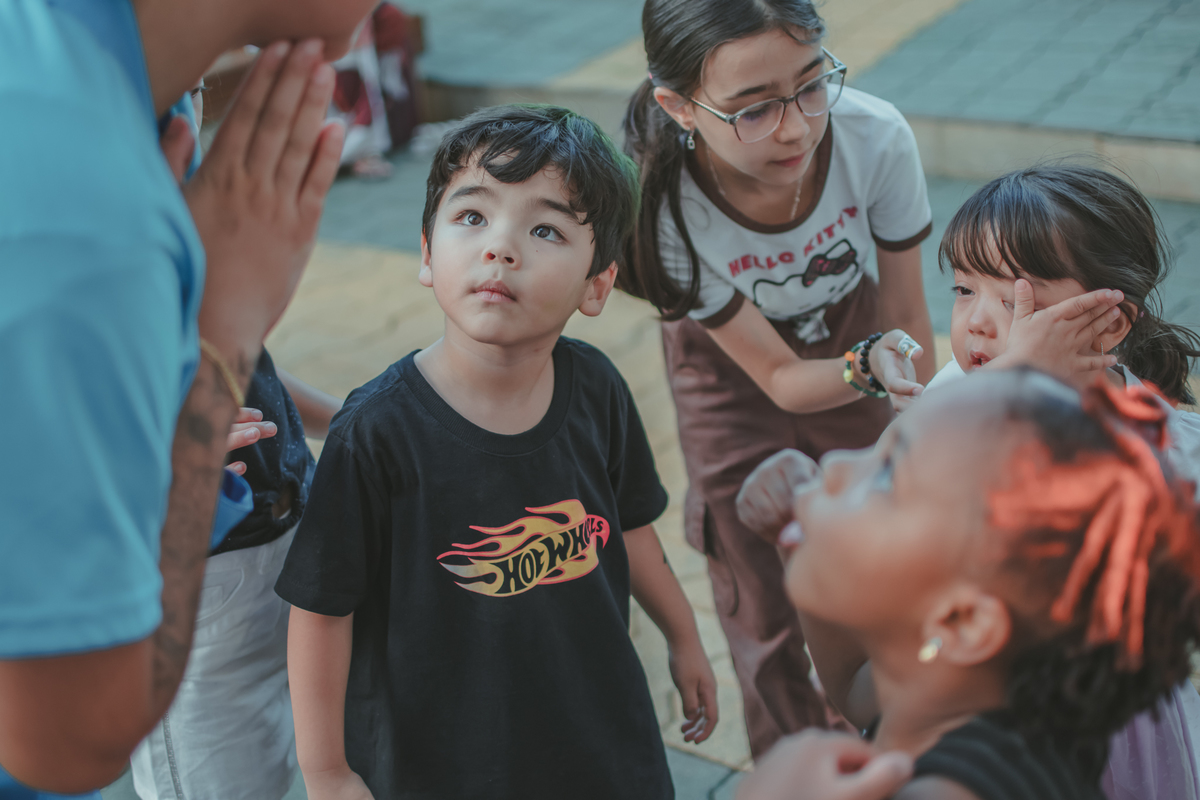 Varias crianças prestando atenção em uma mulher dando orientações para brincadeiras, durante uma festa de aniversário infantil.
