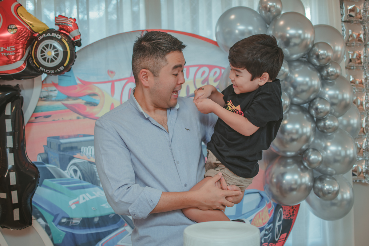 Um homem surpreso com o desenho na mão do filho, os dois em frente uma decoração de aniversário com tema Hot Wheels.