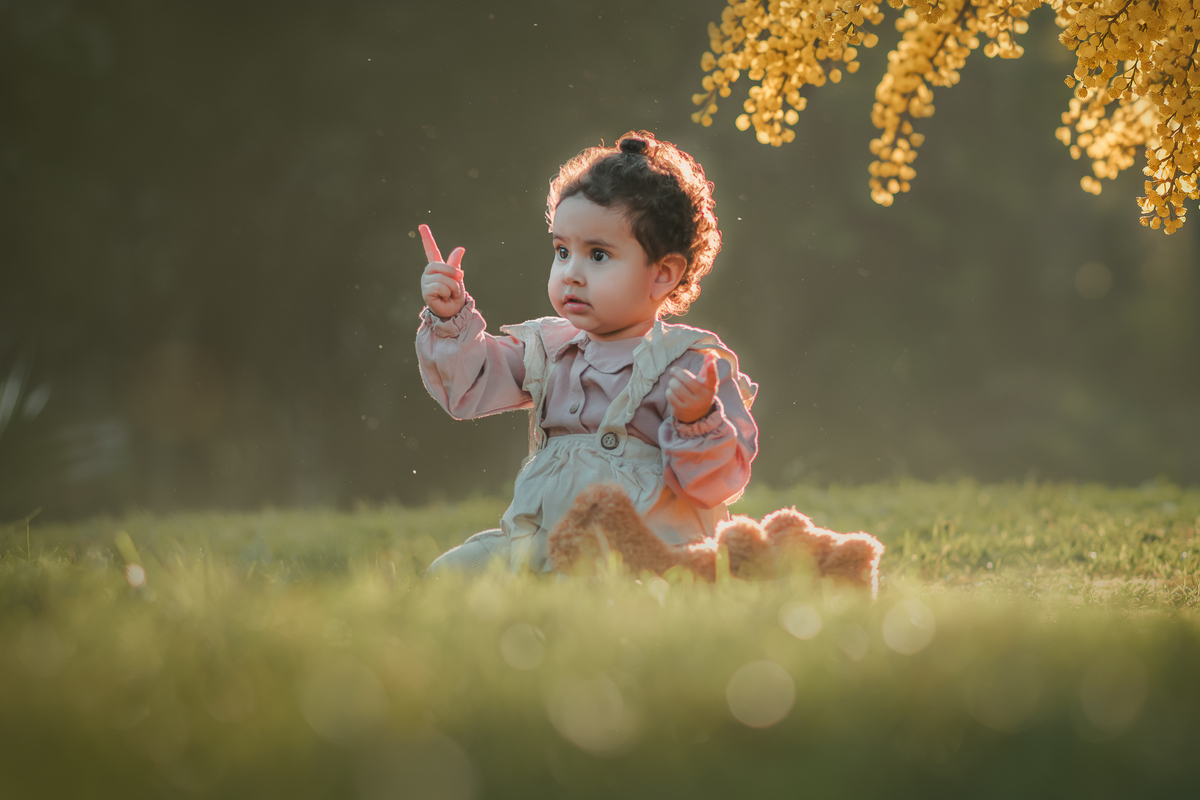 Uma bebê vestidda com roupas camponesa sentada na grama e brincando com um urso de pelúcia, durante seu ensaio de 1 ano.