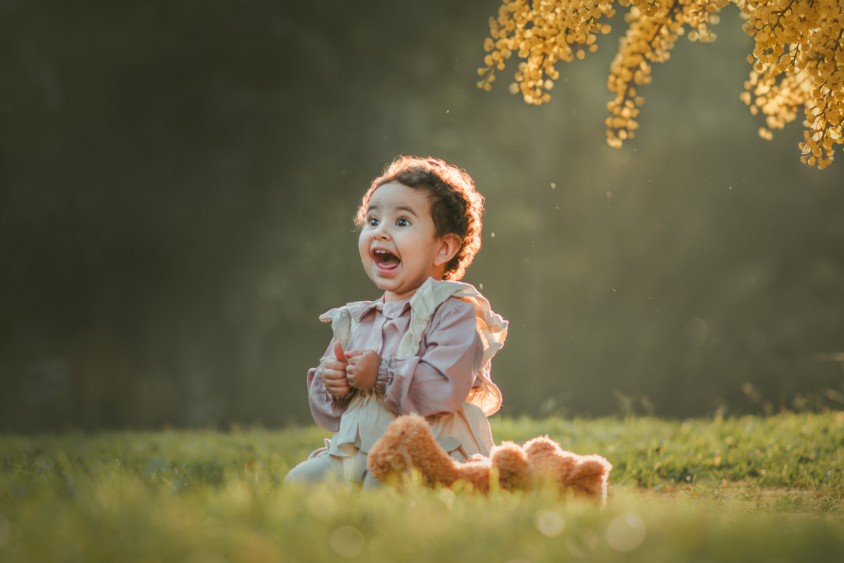 Uma bebê vestidda com roupas camponesa sentada na grama e brincando com um urso de pelúcia, durante seu ensaio de 1 ano.