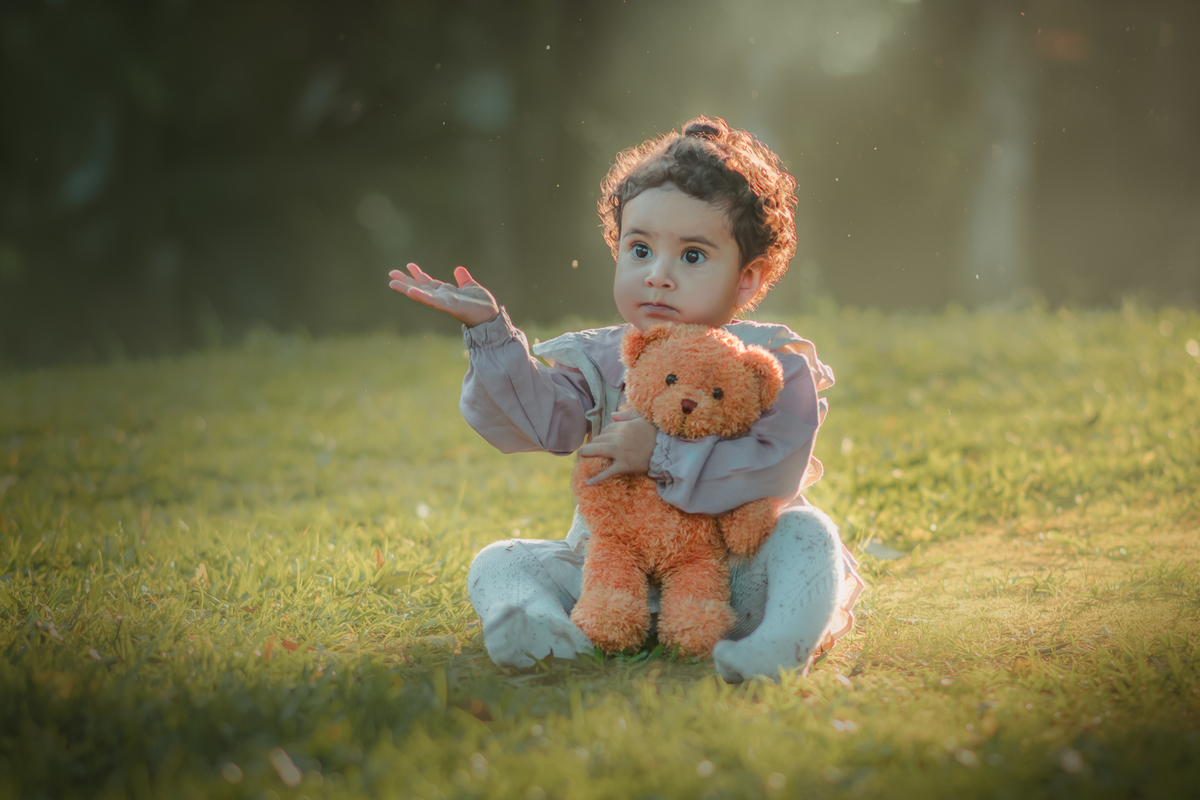 Uma bebê vestidda com roupas camponesa sentada na grama e brincando com um urso de pelúcia, durante seu ensaio de 1 ano.
