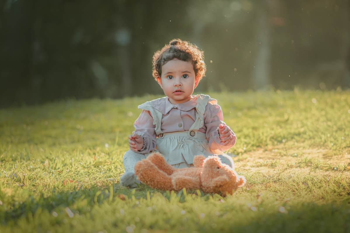Uma bebê vestidda com roupas camponesa sentada na grama e brincando com um urso de pelúcia, durante seu ensaio de 1 ano.