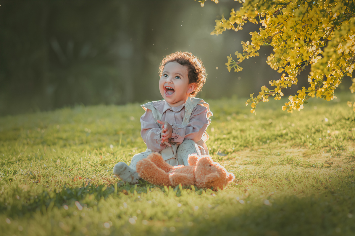 Uma bebê vestidda com roupas camponesa sentada na grama e brincando com um urso de pelúcia, durante seu ensaio de 1 ano.